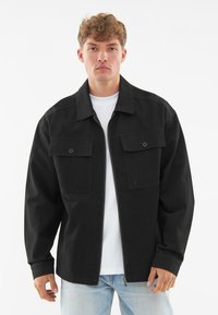 Fekete farmer stílusú dzseki gallérral, két elülső zsebbel és cipzáros záródással. Hosszú ujjak, laza fit. Fehér pólóval kombinálva.