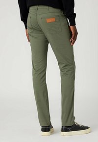 Gröna, raka byxor i slitstark tyg, med en läderlapp på bakfickan och kontrasterande sömnad. Kända med svarta sneakers.