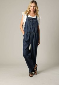 Donkerdenim overall met verstelbare banden, een voorzak, een aangesnoerd taille en brede pijpen. Draagt over een witte top met kant-geborduurde details.