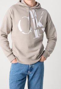 Sudadera beige con gran lettering gráfico en blanco, con capucha ajustable, puños de canalé y un fit relajado, combinada con jeans de mezclilla azules.