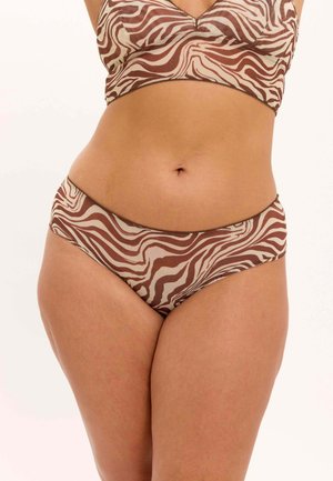 Nainen, joka pukeutunut ruskeaan ja beigeen seepra-raidalliseen rintaliiviin ja siihen sopiviin bikini-tyylisiin alushousuihin yksiväristä vaaleaa taustaa vasten.