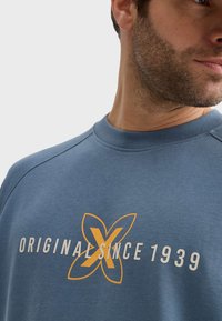 Sudadera azul con cuello redondo y puños acanalados. Presenta un diseño de texto y logo en naranja y blanco que dice "ORIGINAL DESDE 1939".
