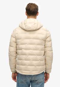 Beige puffjakke med hætte, som har et horisontalt quiltet mønster, elastiske manchetter og en glat overflade. Bagviewet viser et enkelt design.