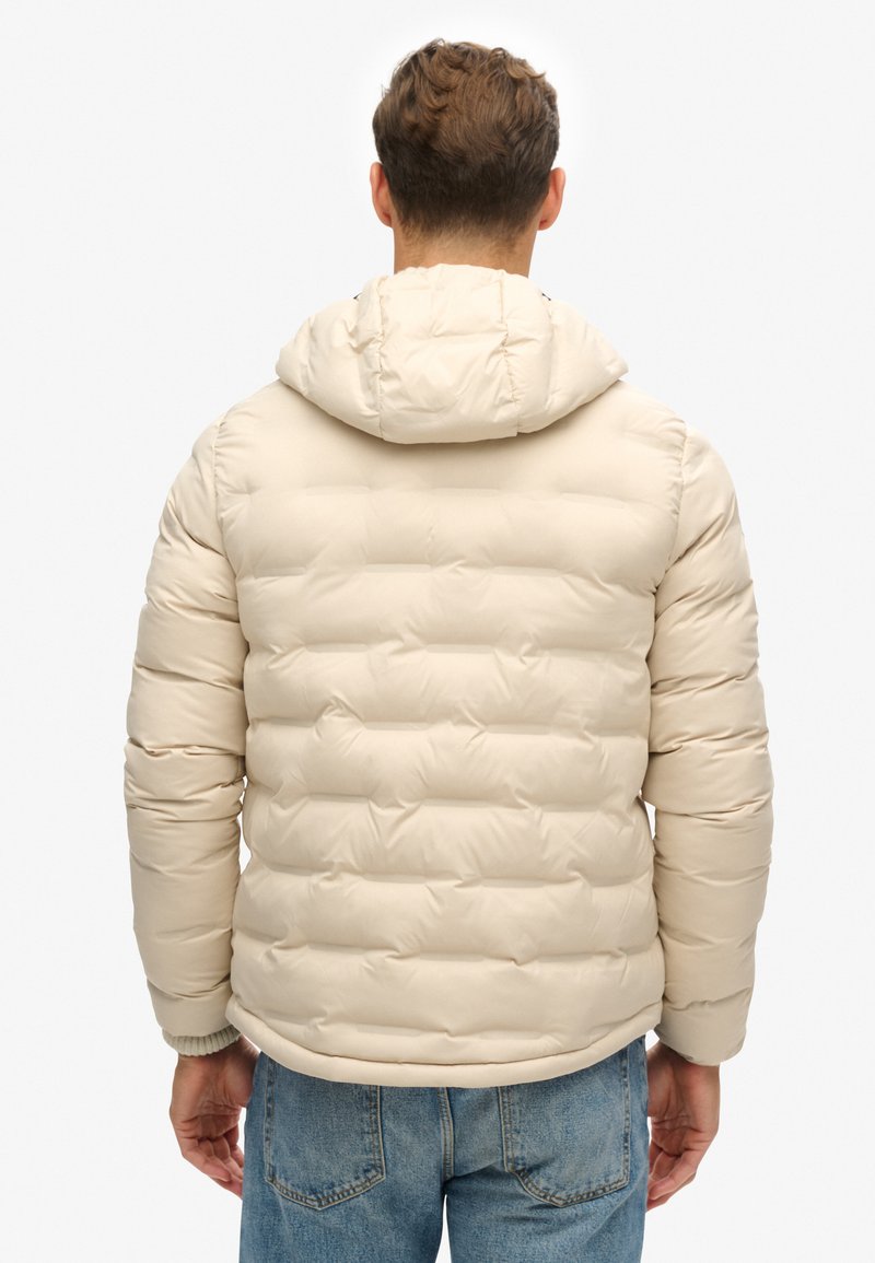 Beige pufferjack met een capuchon, voorzien van een horizontaal gestikt patroon, elastische manchetten en een gladde buitenkant. Achteraanzicht toont een eenvoudig ontwerp.