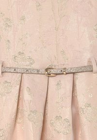 Růžová látka s květinovým vzorem a kovovými nitkami. Stříbrný třpytivý pásek se zlatou sponou zpřístrojí pas, čímž zvýrazňuje texturovaný design.