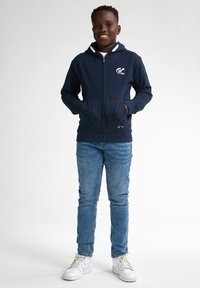 Felpa con zip di colore blu navy con un logo bianco sul petto, dotata di cappuccio foderato, due tasche anteriori e un design aderente, abbinata a jeans chiari e sneakers bianche.