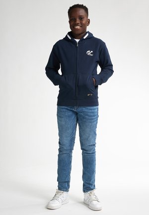 Hoodie zippé bleu marine avec un logo blanc sur la poitrine, dotée d'une capuche doublée, de deux poches avant et d'une coupe ajustée, associée à un jean clair et des baskets blanches.