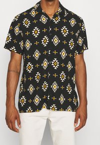 Mężczyzna ubrany w czarną koszulę z krótkim rękawem zapinaną na guziki, z żółtymi i białymi geometrycznymi wzorami w romby, w komplecie z białymi spodniami.