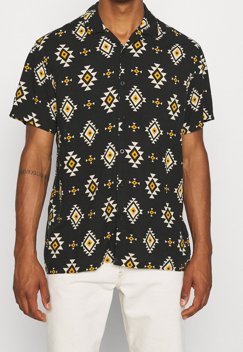 Mężczyzna ubrany w czarną koszulę z krótkim rękawem zapinaną na guziki, z żółtymi i białymi geometrycznymi wzorami w romby, w komplecie z białymi spodniami.