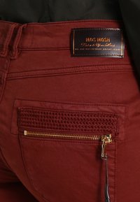 Red jeans with a textured pocket, featuring a gold zipper and a brown leather tag embossed with "MOS MOSH." Smooth fabric finish.