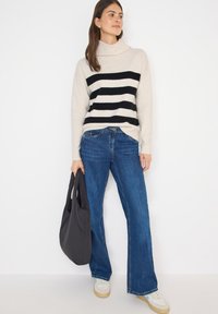 Beige- und schwarz gestreifter Pullover mit Cowl-Neck, kombiniert mit blauen Schlagjeans und weißen Sneakers. Hält eine schwarze Tragetasche.