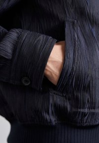 Main dans la poche latérale d'une veste noire texturée avec bouton de manchette visible et ourlet côtelé.