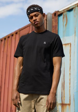 Black cotton t-shirt with short sleeves, featuring a small white embroidered logo on the chest. Model wears khaki shorts and a black bandana.