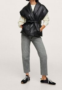 Chaleco de cuero negro con solapas anchas y cintura ceñida; combinado con jeans rectos grises y zapatillas negras con cordones blancos.