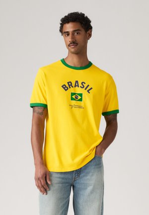 Muž s kudrnatými vlasy a knírem, který má na sobě žluté tričko se zeleným lemováním a brazilskou vlajkou, doplněné světle modrými džíny, stojí na jednobarevném pozadí.