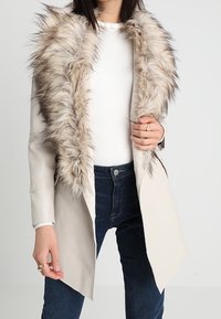 Abrigo largo beige con un cuello de piel sintética de color marrón claro. El abrigo presenta un corte ajustado y un diseño elegante, con un brillo sutil y un hardware mínimo.