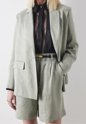 Frau trägt hellgrünen strukturierten Blazer und passende Shorts über einer transparenten schwarzen Hemdbluse mit Kragen und schwarzem Gürtel mit goldener Schnalle.