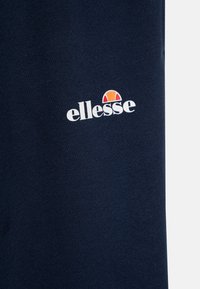 Jūras zilas kokvilnas audums ar teksturētu virsmu, ar balto "ellesse" logo un krāsainu pusapaļu akcentu virs tā.
