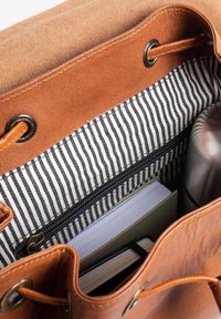 Brown leather bag with striped fabric lining, containing a closed notebook, a book, and a stainless steel travel mug inside.