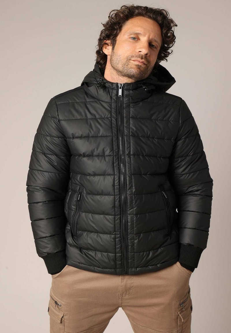 Doudoune noire avec un design ajusté, piqûres horizontales, capuche, zip avant et poches latérales. Équipée de poignets côtelés pour plus de chaleur.