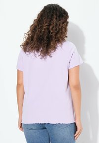 Top morado de manga corta con dobladillo en forma de onda y textura suave, combinado con jeans azul claro, mostrado desde la parte trasera sobre un fondo blanco.