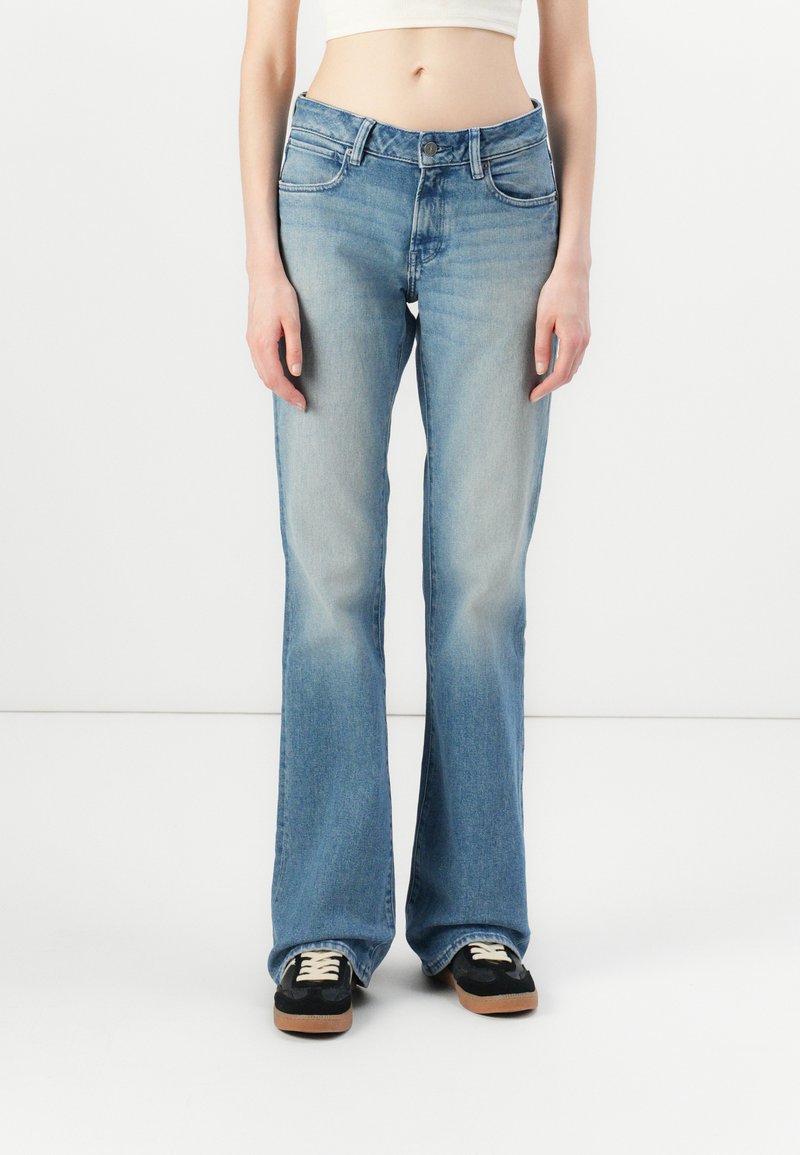 Jeans evazați bleu deschis din denim cu un finisaj decolorat, design cu cinci buzunare și bucle pentru curea, asortați cu teniși negri și un top crem.
