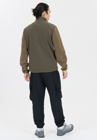 Fleece jacket with olive green body and textured brown sleeves, paired with black cargo pants and gray sneakers, viewed from the back.