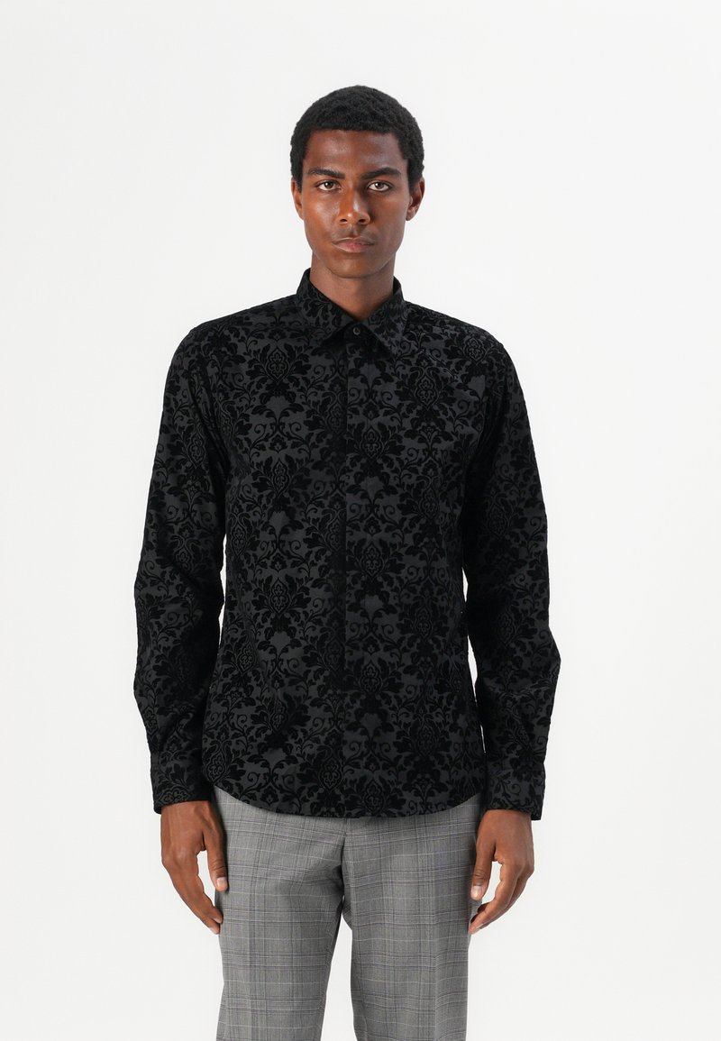 Black long-sleeve shirt with a textured floral pattern, collared design, and button front. Worn with gray plaid trousers.