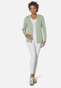 Hellgrüner, gerippter Cardigan aus weichem Material, mit V-Ausschnitt, langen Ärmeln und Knöpfen, kombiniert mit weißen Hosen und Sneakern.