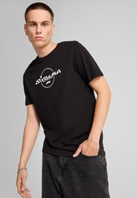 Czarny bawełniany t-shirt z białym graficznym logo z przodu. Model ma na sobie ciemne dżinsy o luźnym kroju i widocznej teksturze.