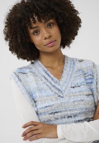 Fiatal nő göndör hajjal, kék és szürke kötött mellényt visel fehér, hosszú ujjú póló felett, karjai keresztbe tett állapotban, semleges háttérrel.