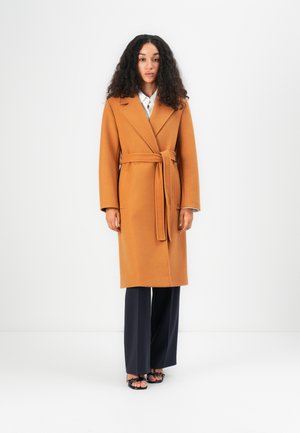 Long, wrap-style coat in warm orange, with a wide collar, belt at the waist, and long sleeves; worn over dark trousers.