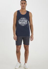 Mørkeblå tanktop med hvid grafisk tryk, parret med mørkegrå shorts. Modellen har hvide sneakers på; ensfarvet baggrund.