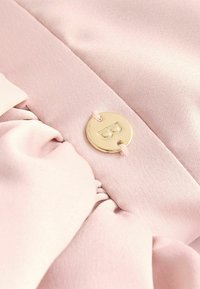 Pūķa rozā audums ar savilkta tekstūru un zelta toņa apaļu akcentu, nostiprinātu ar atbilstošu auklu, uz kuras ir attēlota burts "B".