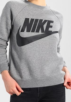 Persona vistiendo una sudadera gris de Nike con un gran logo negro de Nike y el símbolo swoosh en el frente, con las manos descansando cerca de las caderas.