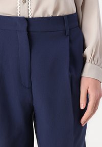 Marineblå bukser med en glat tekstur, der har en flat front, sidelommer og en mellemhøj talje. Hånd placeret på stoffet.