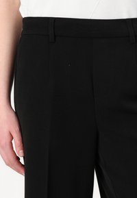 Pantaloni negri croiți, cu o textură netedă, având picior drept, curelușe pentru centură și buzunare laterale, prezentati la jumatatea tivului cu o mână.