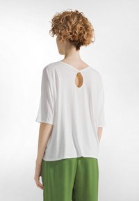 Osoba o krótkich kręconych włosach ubrana w luźną białą bluzkę z małym wycięciem w kształcie klucza na górnej części pleców oraz zielone spodnie, stojąca na białym tle.