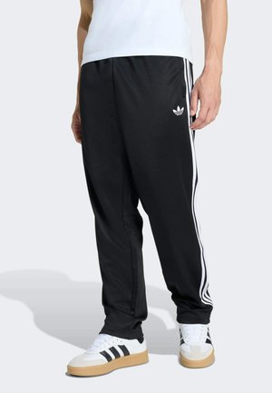 Person trägt schwarze Adidas-Trainingshose mit weißen Streifen und Logo, kombiniert mit weißen Sneakers mit schwarzen Streifen und beigefarbenen Sohlen.