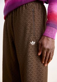 Bruna joggers med textur och ett subtilt mönster, sidofickor med dragkedja och vit Adidas-logotyp nära midjan. Tillsammans med en flerfärgad tröja.