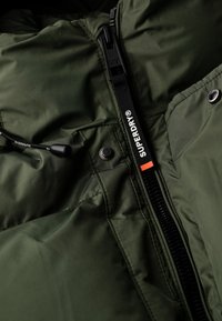 Zbliżenie na zamek błyskawiczny i zatrzask zielonej kurtki Superdry z markowym uchwytem oraz detalem czarnej linki.