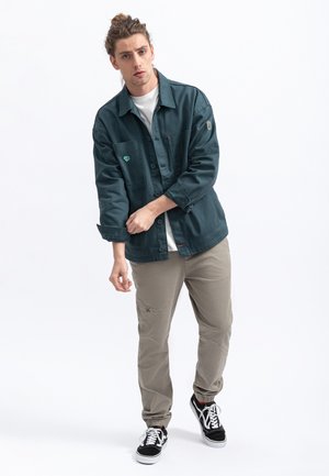 Dark teal jacket with button closure and pockets, paired with light khaki tapered pants and black sneakers with white laces.