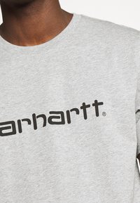 Close-up van een persoon in een lichtgrijs T-shirt met zwarte "Carhartt" logo-tekst over de borst.