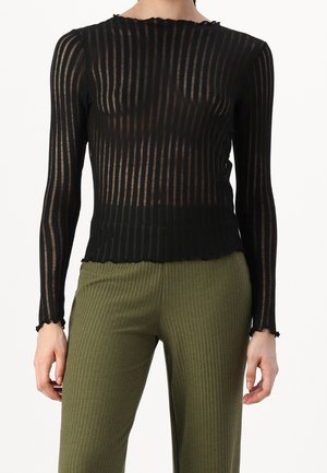 Top nero trasparente a maniche lunghe con righe verticali abbinato a pantaloni verdi oliva a costine, indossato da una persona in piedi su sfondo bianco.