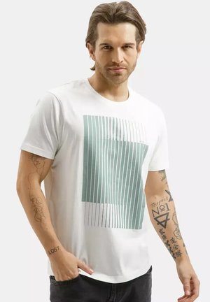 Mężczyzna z jasnobrązowymi włosami ubrany w białą koszulkę z turkusowymi geometrycznymi paskami, pokazujący tatuaże na obu przedramionach, stojący na białym tle.