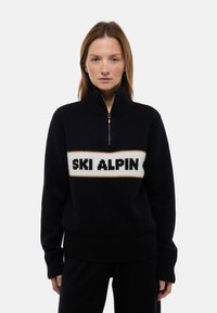 Pull noir avec un col montant, dotée d'une fermeture éclair et d'un bandeau central blanc avec "SKI ALPIN" en texte noir. Texture en maille douce.