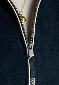 Gros plan sur une veste bleu marine foncé partiellement zippée montrant un curseur de fermeture éclair argenté et un collier pendentif doré avec un motif de visage sous la fermeture éclair.