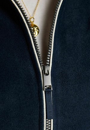 Close-up af en delvist lynet mørkeblå jakke, der viser en sølv lynlåstrækker og en guldfarvet vedhængshalskæde med ansigtsdesign under lynlåsen.