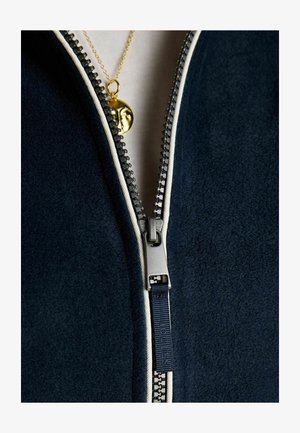 Záber zblízka na čiastočne zapnutú tmavomodrú bundu so strieborným zipsom a zlatým príveskom na náhrdelníku s motívom tváre pod zipsom.