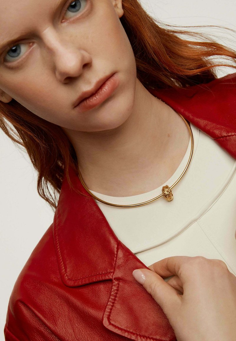 Collar de oro con un acento decorativo, llevado sobre una blusa blanca, complementado por una chaqueta de cuero roja con detalles de costura prominentes.